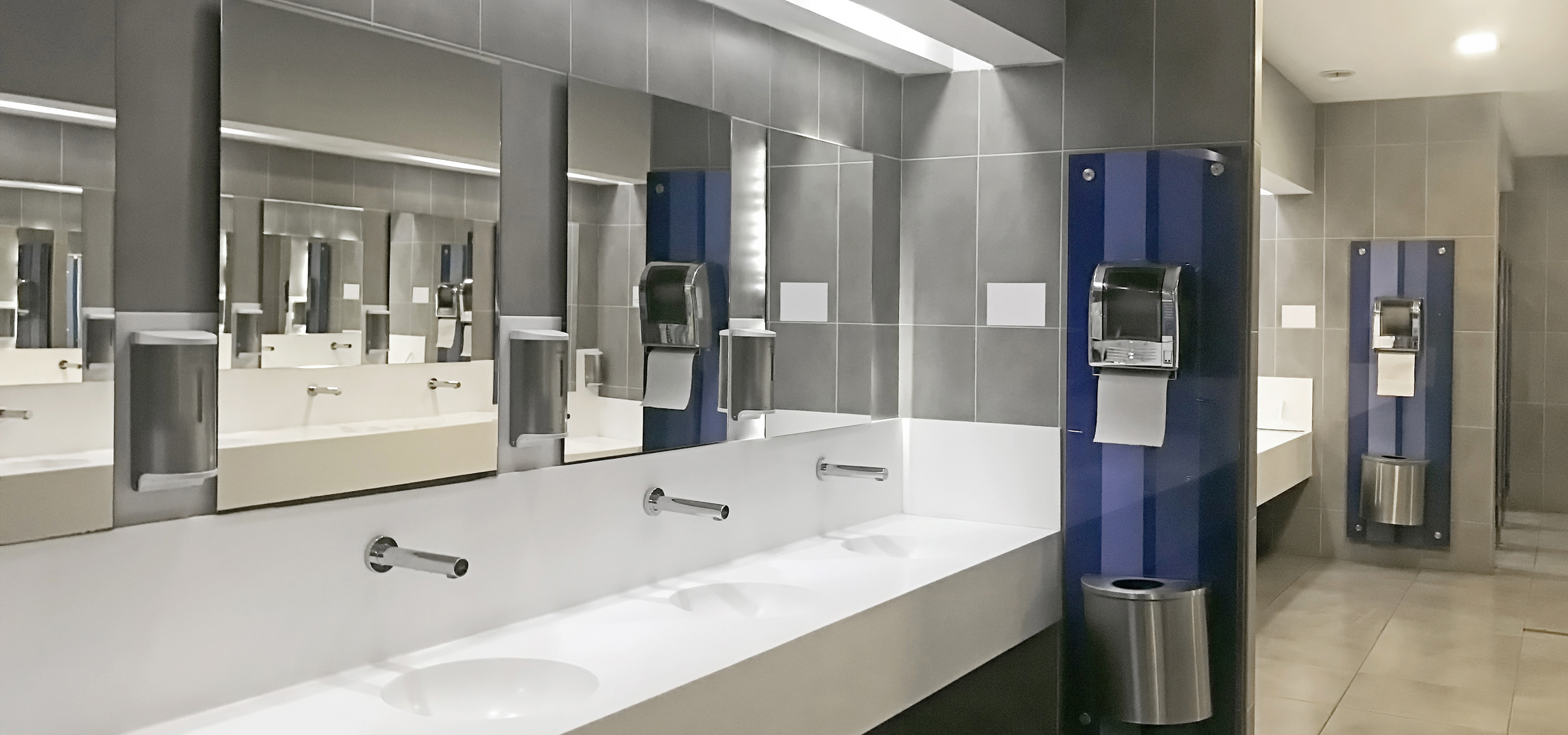 Wide angle, perspective view of public restroom in which the mirrors shown reflect mirrors on the opposite side creating an optical illusion. The image contains many commercial bathroom accessories such as soap dispensers, mirrors, and paper towel dispensers with built in waste receptacles.