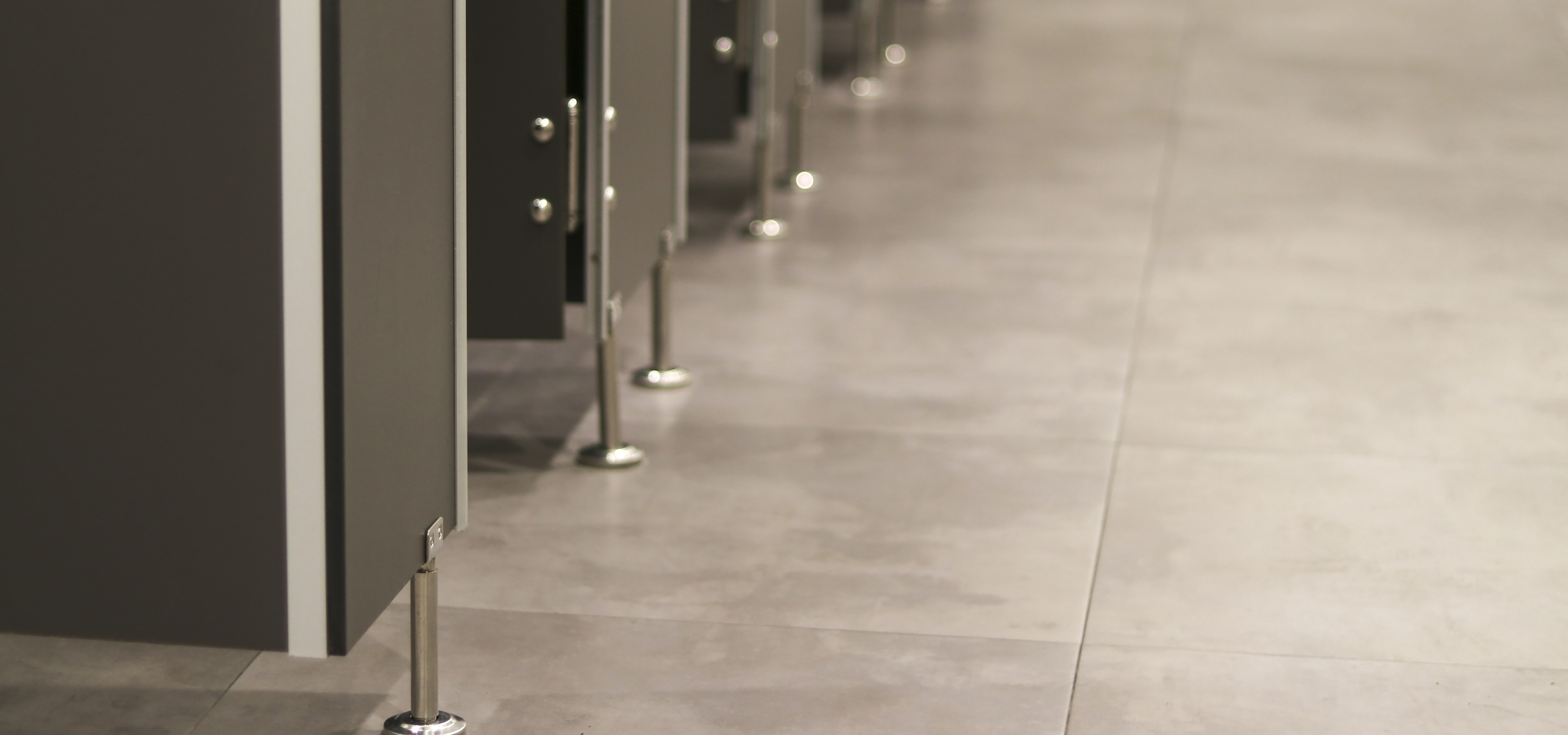A shot close to the floor of a public bathroom, creating a perspective effect in which hardware for floor-mounted partitions are shown sweeping up from lower left to top center on one side, and the other side is the tile floor upon which they rest.